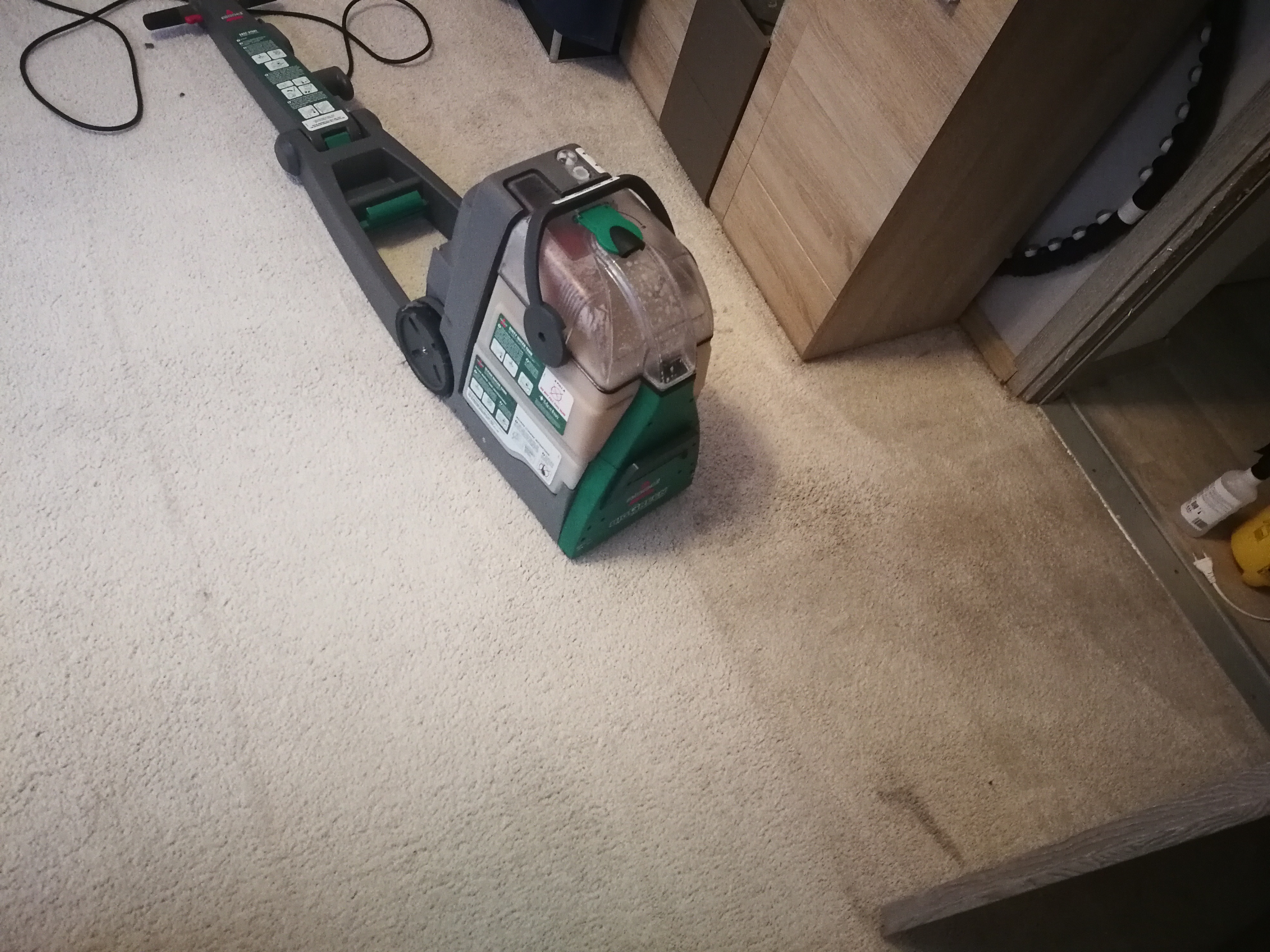 Maszyna do czyszczenia dywanów marki Bissell na jasnym dywanie, widoczny zbiornik z brudną wodą, obok szafka z drewna.