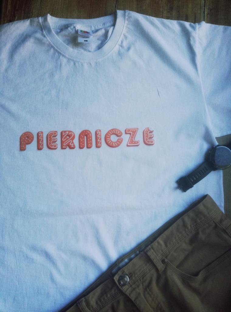 Biały t-shirt z napisem 'PIERNICZE' stylizowanym na pierniki, leżący obok brązowych spodni i czarnego smartwatcha na drewnianym podłożu.
