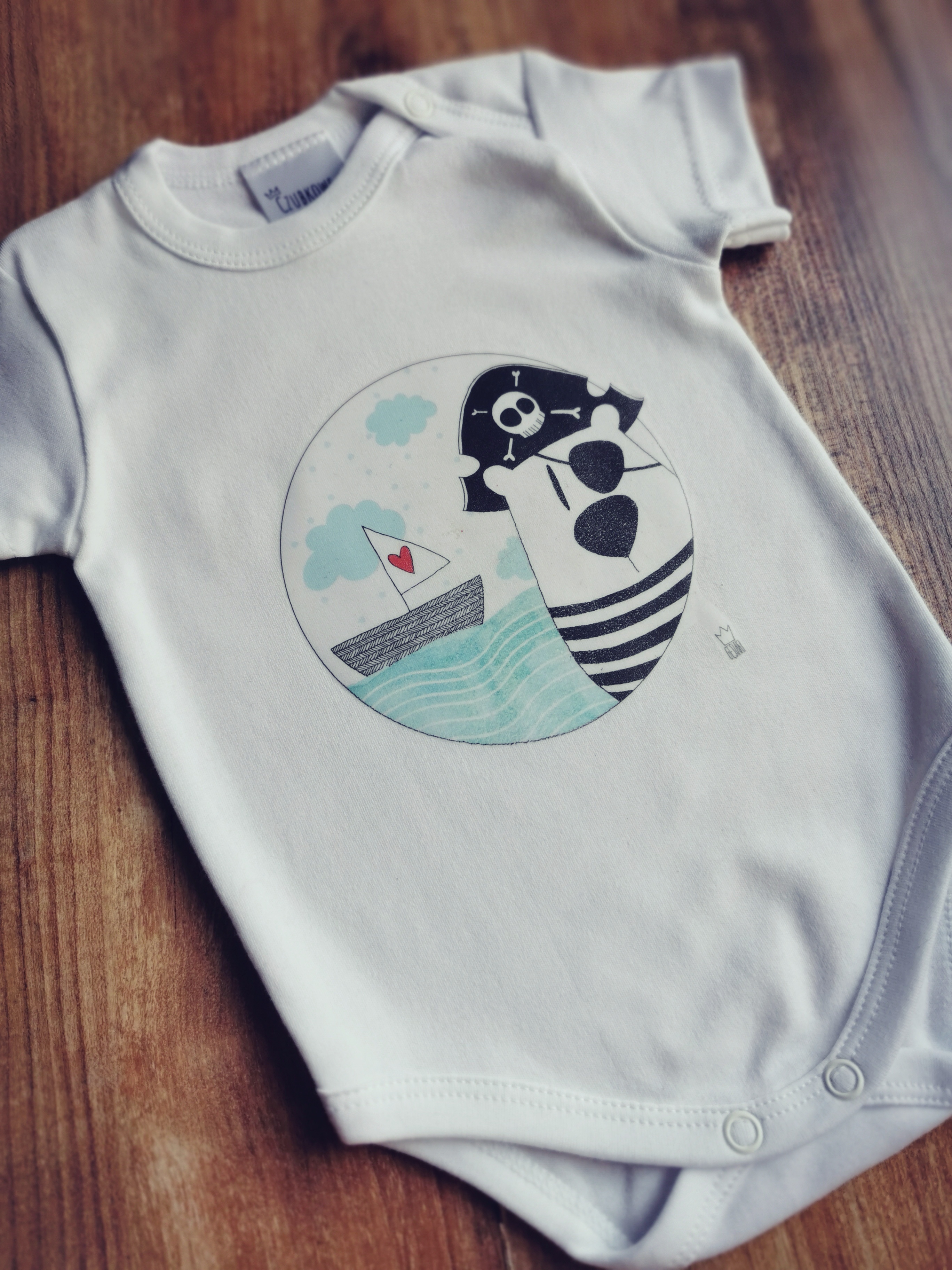 Białe niemowlęce body z krótkim rękawem, ozdobione graficznym nadrukiem przedstawiającym pirata w okularach i łódkę z sercem na żaglu, leżące na drewnianym podłożu.