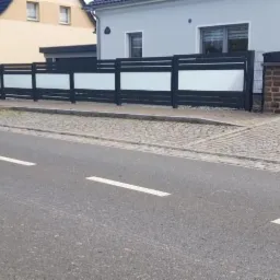 Nowoczesne ogrodzenie z poziomych, grafitowych paneli z mlecznym szkłem, oddzielające posesję od ulicy z kostki brukowej i asfaltowej drogi.