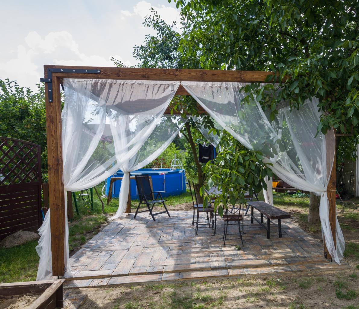 Drewniana altana z białymi zasłonami na kamiennej podłodze, z widokiem na basen w ogrodzie.