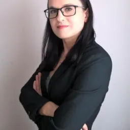 Portret kobiety w okularach i czarnej marynarce z założonymi rękami na neutralnym tle.