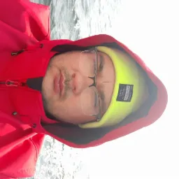 Portret mężczyzny w czerwonej kurtce z kapturem i neonowożółtej czapce z napisem STREETSTYLE, na tle wody.