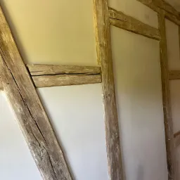 Fragment ściany z drewnianą konstrukcją szkieletową, częściowo pokrytej jasną, gładką warstwą wykończeniową, prawdopodobnie świeżo położoną.