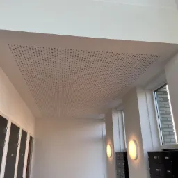 Biały sufit podwieszany z perforacją w regularne wzory, widok z dołu w korytarzu z szafkami i oświetleniem ściennym.