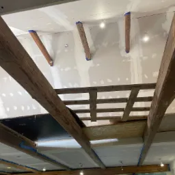 Montaż sufitu podwieszanego z widocznymi belkami drewnianymi, zabezpieczonymi niebieską taśmą malarską, oraz fragmentami konstrukcji nośnej i oświetleniem punktowym.