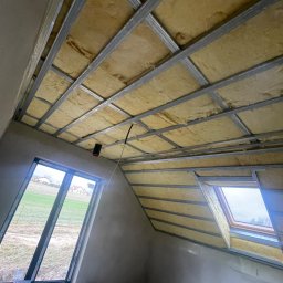 Słonina Klusbedrijf Ireneusz Słonina - Poddasze w trakcie ocieplania: widok na konstrukcję metalową z wypełnieniem izolacyjnym, okno dachowe i okno pionowe z widokiem na zewnątrz.