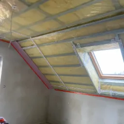 Poddasze w trakcie ocieplania, widoczna konstrukcja metalowa, żółta izolacja zabezpieczona folią, różowe panele izolacyjne przy ścianie oraz okno dachowe z taśmami uszczelniającymi.