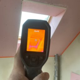 Słonina Klusbedrijf Ireneusz Słonina - Termowizja poddasza w Nowogrodźcu, ukazująca temperaturę 13.4°C na zamontowanej płycie przy oknie dachowym, z widocznymi śladami po wkrętach.