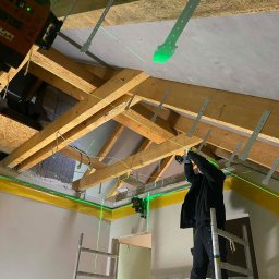 Słonina Klusbedrijf Ireneusz Słonina - Montaż stelaża pod zabudowę poddasza z użyciem poziomicy laserowej Hilti. Widoczne drewniane krokwie i metalowe wieszaki. Praca na wysokości z drabiny.