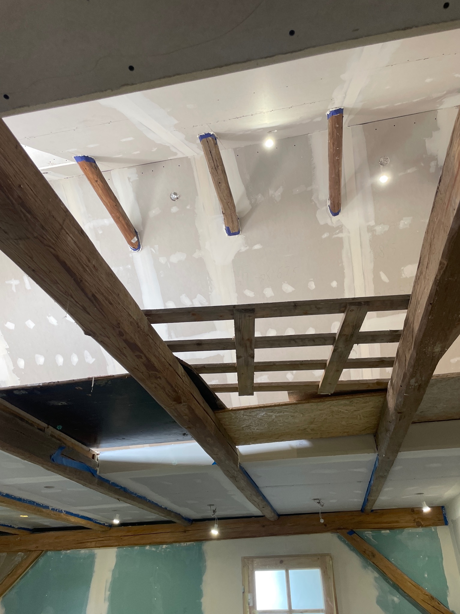 Montaż sufitu podwieszanego z widocznymi belkami drewnianymi, zabezpieczonymi niebieską taśmą malarską, oraz fragmentami konstrukcji nośnej i oświetleniem punktowym.