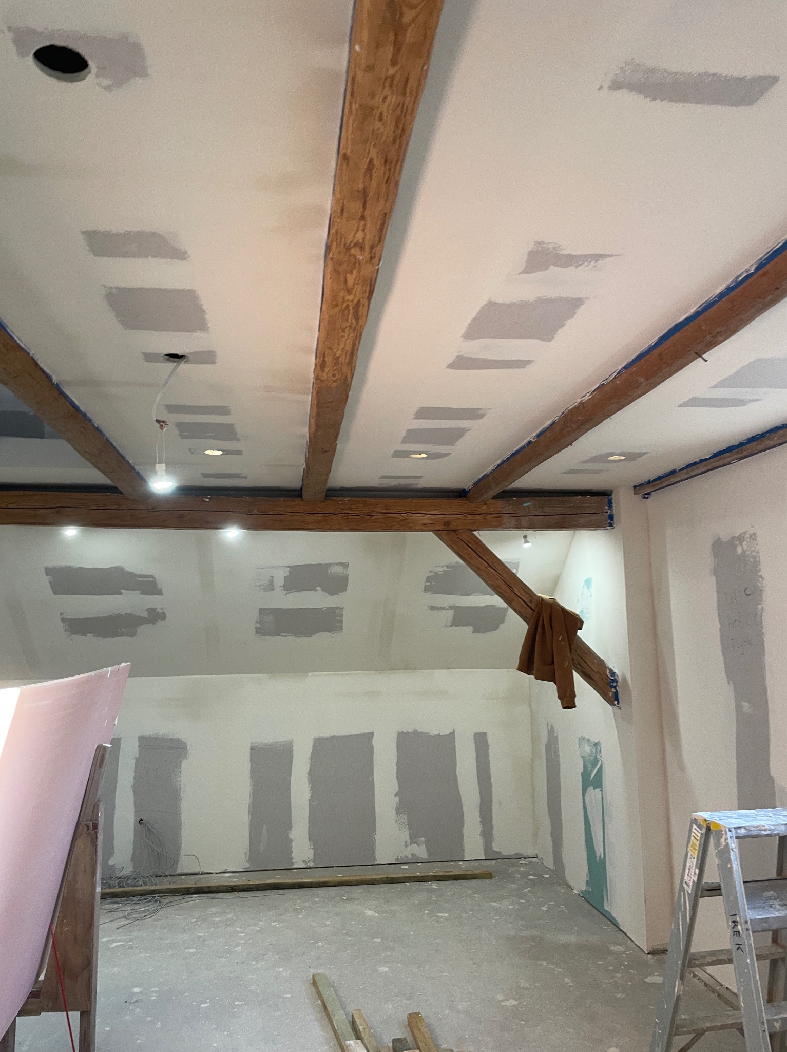 Poddasze w trakcie remontu: drewniane belki stropowe, częściowo zamontowane płyty na suficie i ścianach, widoczne miejsca po szpachlowaniu, wystające kable od oświetlenia, drabina w rogu...