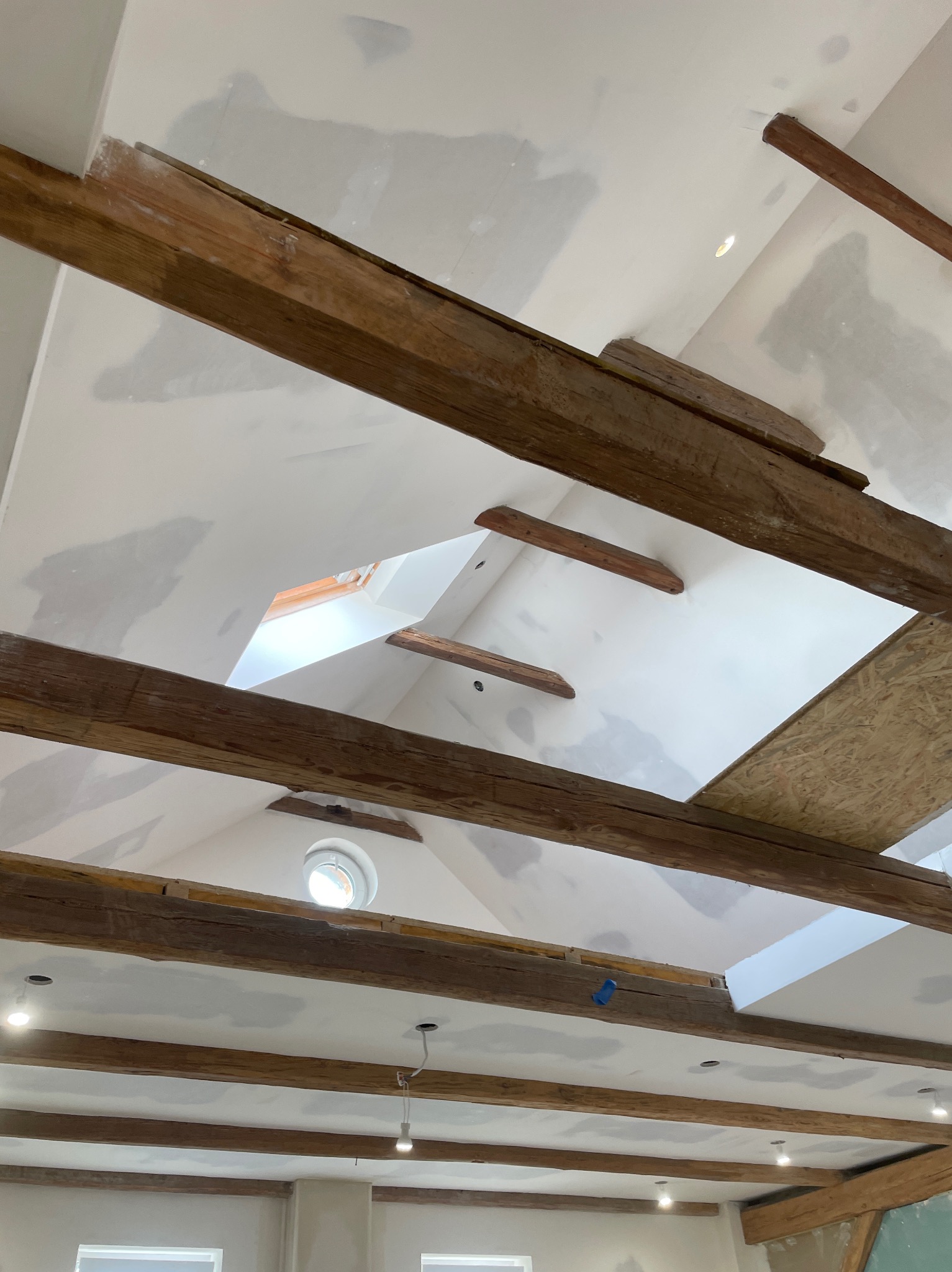 Sufit poddasza z drewnianymi belkami i oknem dachowym w trakcie szpachlowania, widoczne niedoskonałości i przygotowanie do malowania, oświetlenie punktowe.