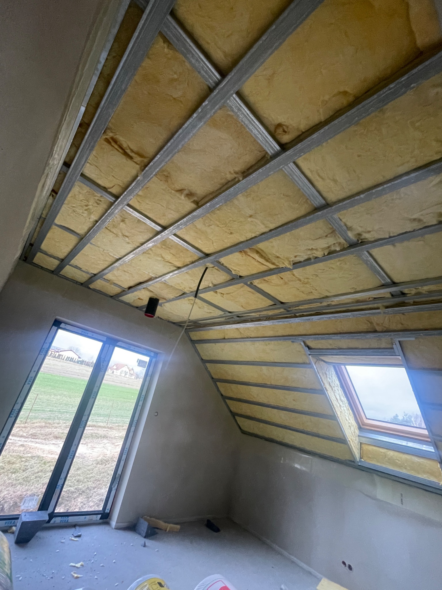 Poddasze w trakcie ocieplania: widok na konstrukcję metalową z wypełnieniem izolacyjnym, okno dachowe i okno pionowe z widokiem na zewnątrz.