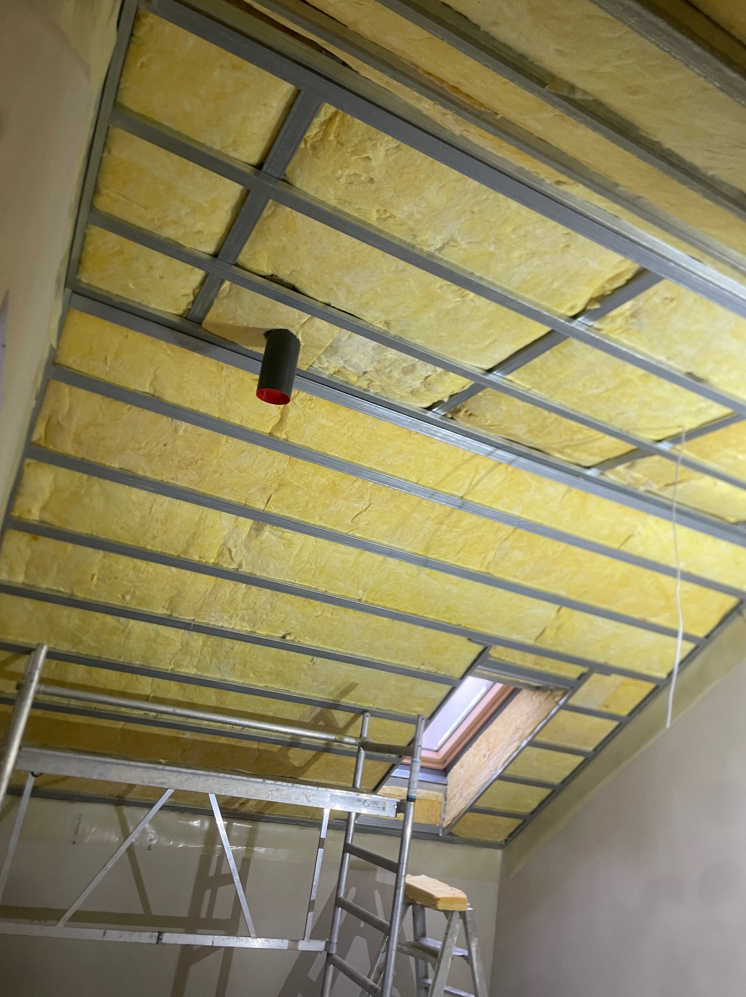 Ocieplone poddasze z widoczną konstrukcją metalową, wełną mineralną między profilami i oknem dachowym, widziane z poziomu rusztowania.