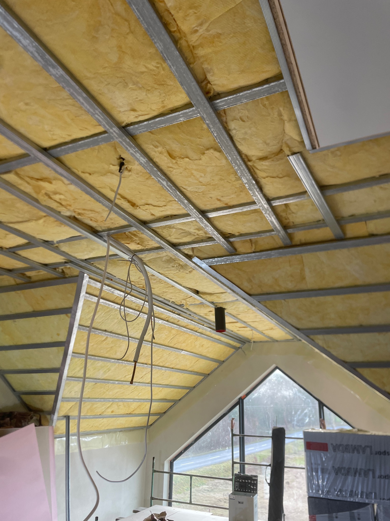 Poddasze w trakcie ocieplania, widoczna konstrukcja z profili stalowych wypełniona żółtą izolacją, przewody elektryczne zwisają z sufitu, okno dachowe w tle.
