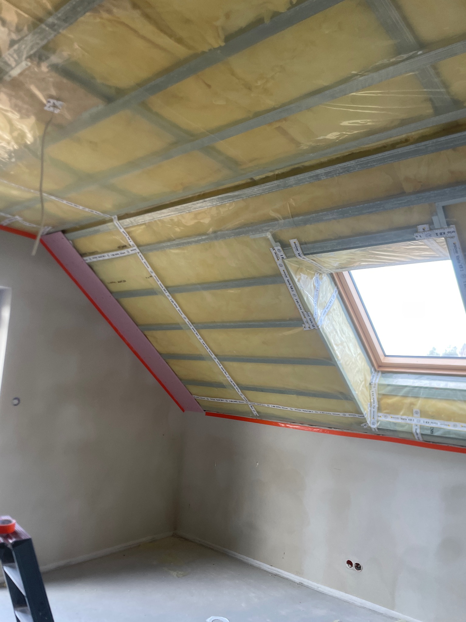 Poddasze w trakcie ocieplania, widoczna konstrukcja metalowa, żółta izolacja zabezpieczona folią, różowe panele izolacyjne przy ścianie oraz okno dachowe z taśmami uszczelniającymi.