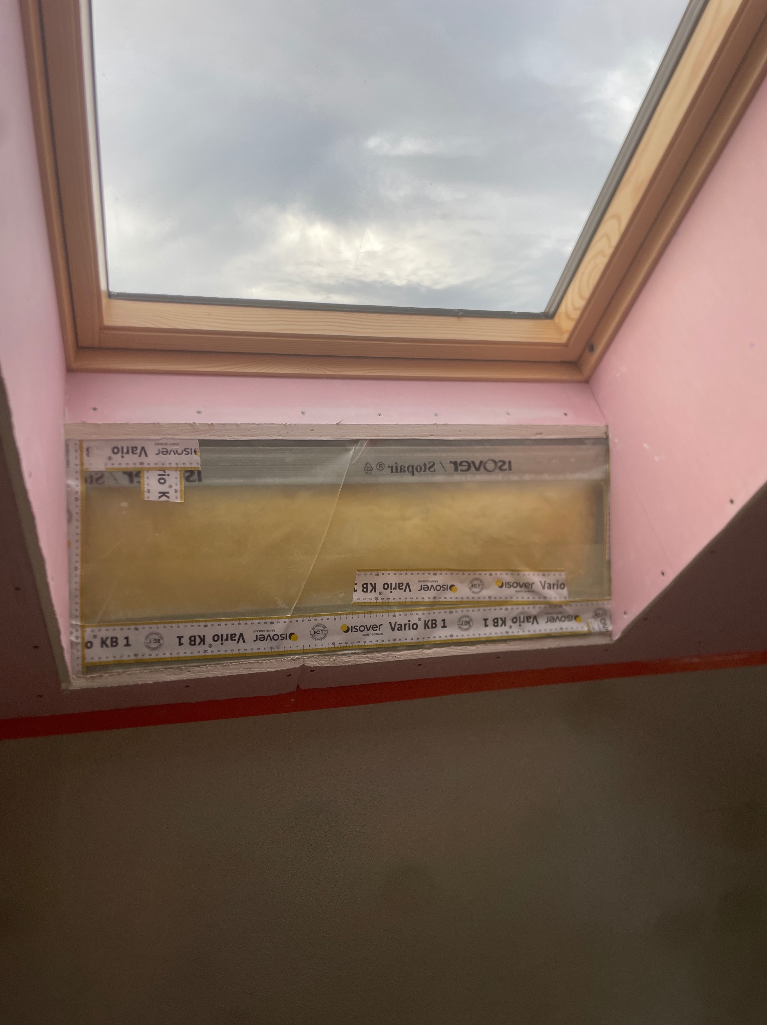 Montaż izolacji z folią paroizolacyjną wokół okna dachowego, widoczne elementy konstrukcyjne poddasza i fragment ściany z płytami gipsowo-kartonowymi.