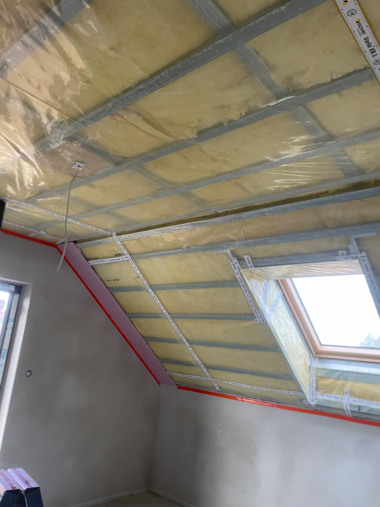 Poddasze w trakcie izolacji: widoczna żółta warstwa izolacyjna, metalowy stelaż, folia paroizolacyjna i okno dachowe z taśmami uszczelniającymi.