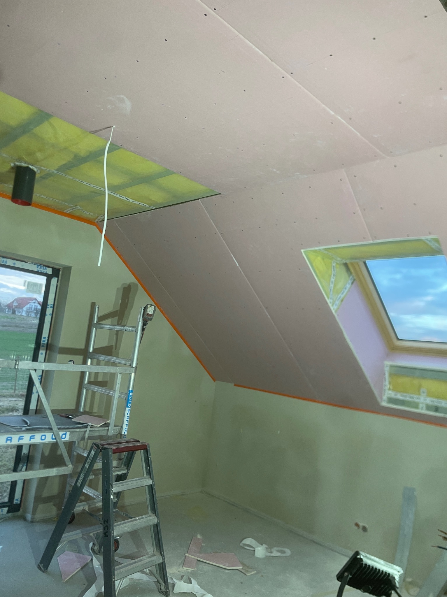 Poddasze w trakcie wykańczania: widoczne płyty gipsowo-kartonowe na skosach i suficie, izolacja wokół okna dachowego, drabiny i przewód elektryczny zwisający z sufitu.