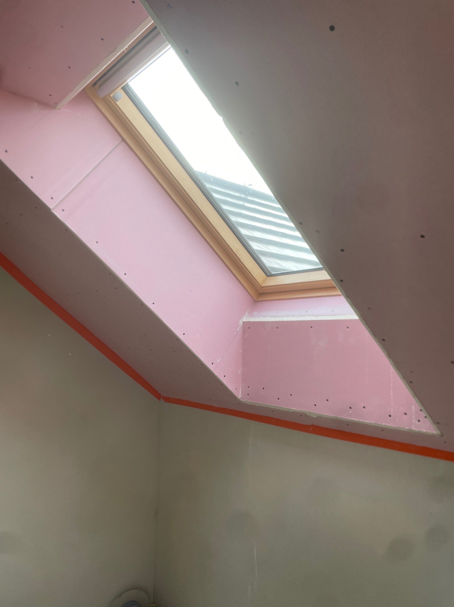 Montaż różowej płyty gipsowo-kartonowej wokół okna dachowego, widoczne wkręty i pomarańczowa taśma malarska na styku ze ścianą.