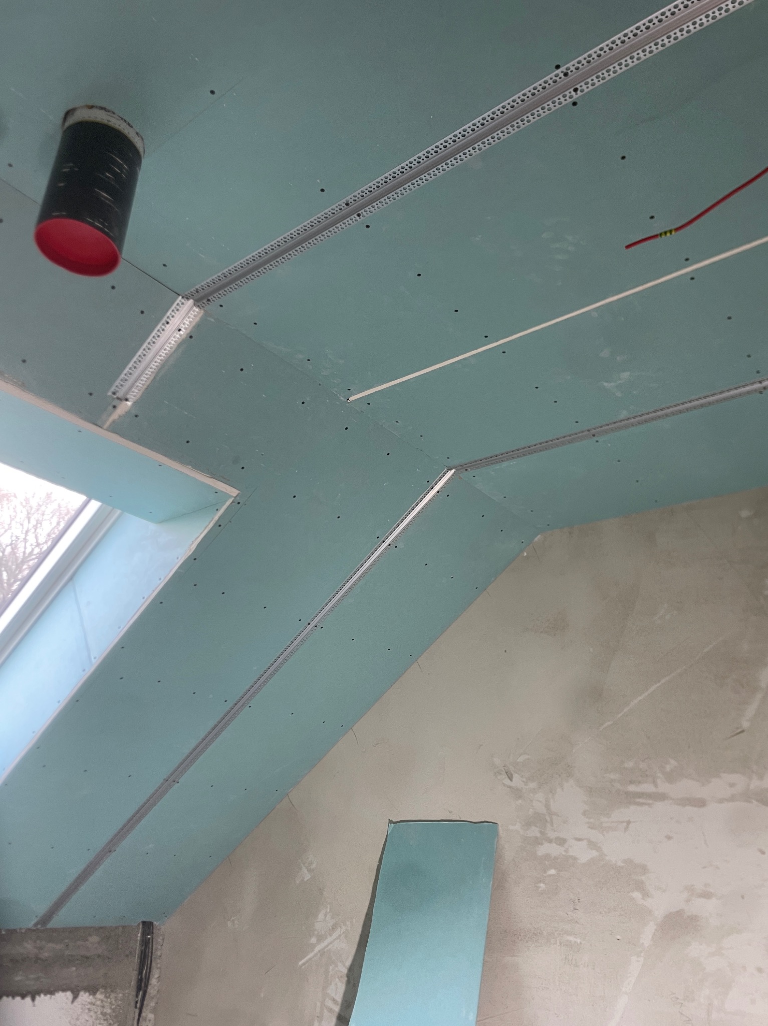 Montaż sufitu podwieszanego z płyt gipsowo-kartonowych, widoczne profile metalowe, otwór wentylacyjny z czerwonym elementem, instalacja elektryczna. Ściany surowe, w trakcie prac wykończeniowych.