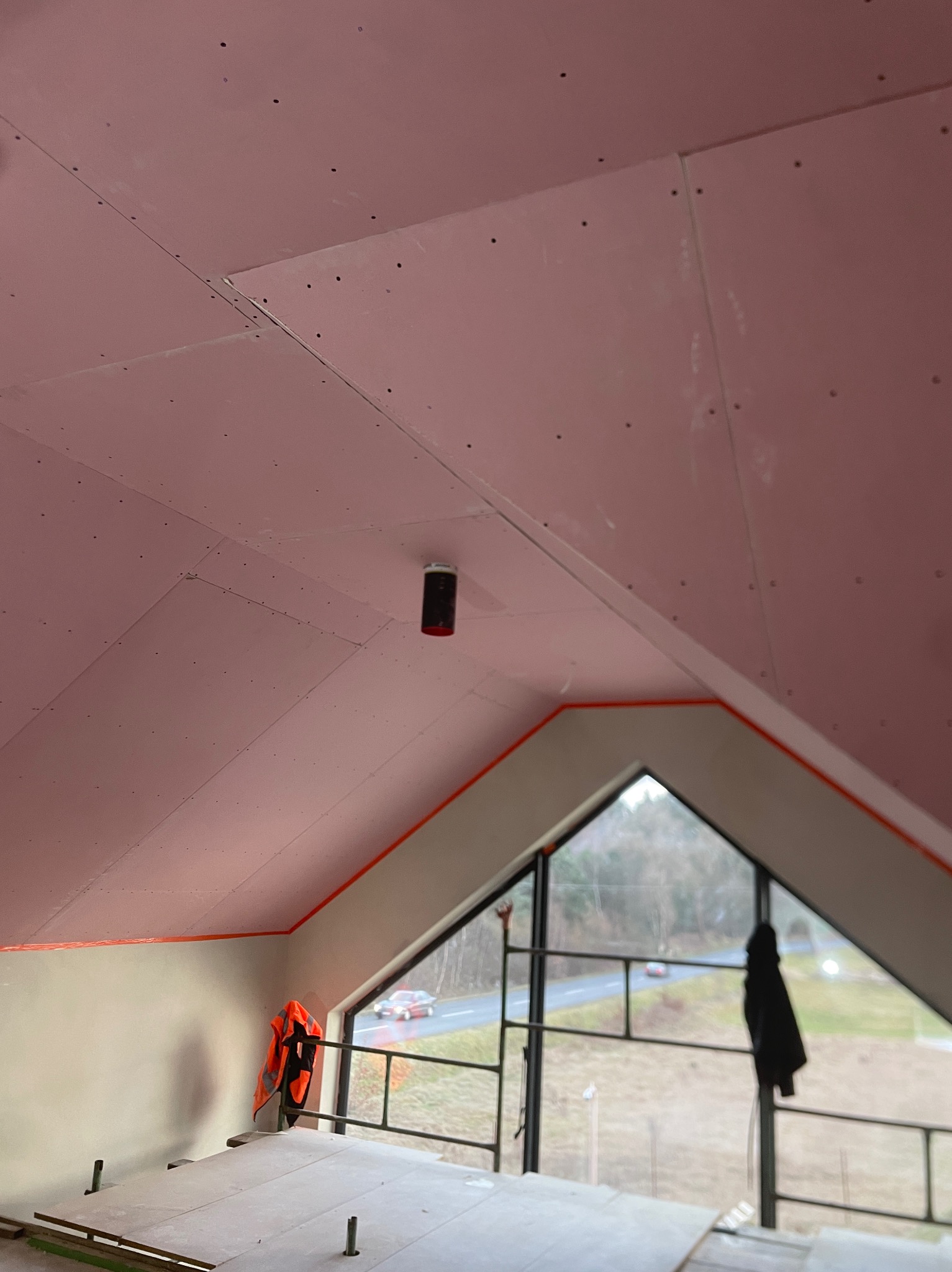 Montaż różowej płyty karton gips na skośnym poddaszu z widocznymi łączeniami, zabezpieczonymi taśmą, oraz fragment rusztowania i wisząca kamizelka odblaskowa.