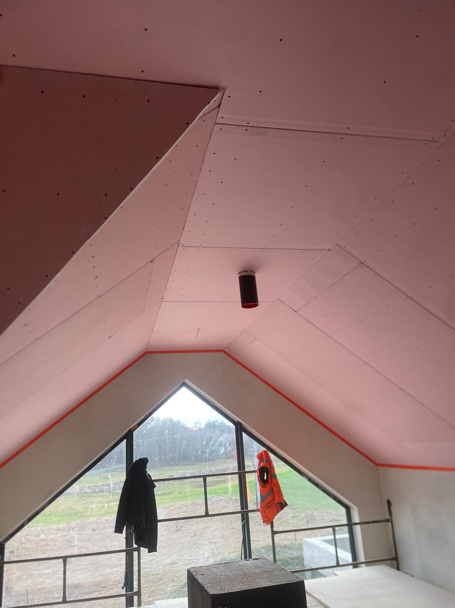 Poddasze z ukończonymi pracami montażu płyt gipsowo-kartonowych, widoczne łączenia i otwory po wkrętach, na oknie wisi kurtka i kamizelka odblaskowa.