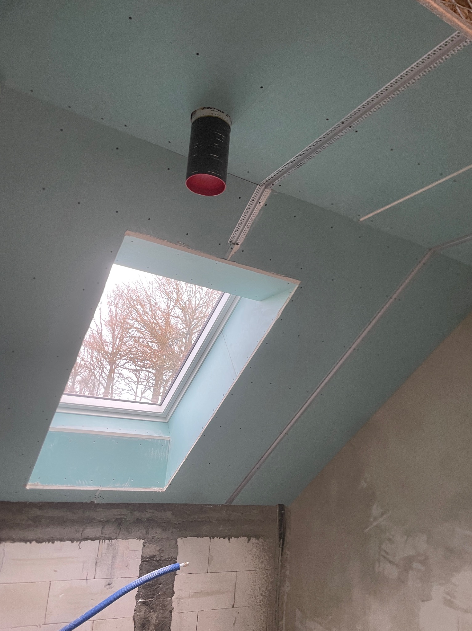 Montaż sufitu poddasza z oknem dachowym, widoczne płyty gipsowo-kartonowe, profil metalowy, otwór wentylacyjny z czarną rurą i czerwoną krawędzią oraz fragment ściany z pustaków.