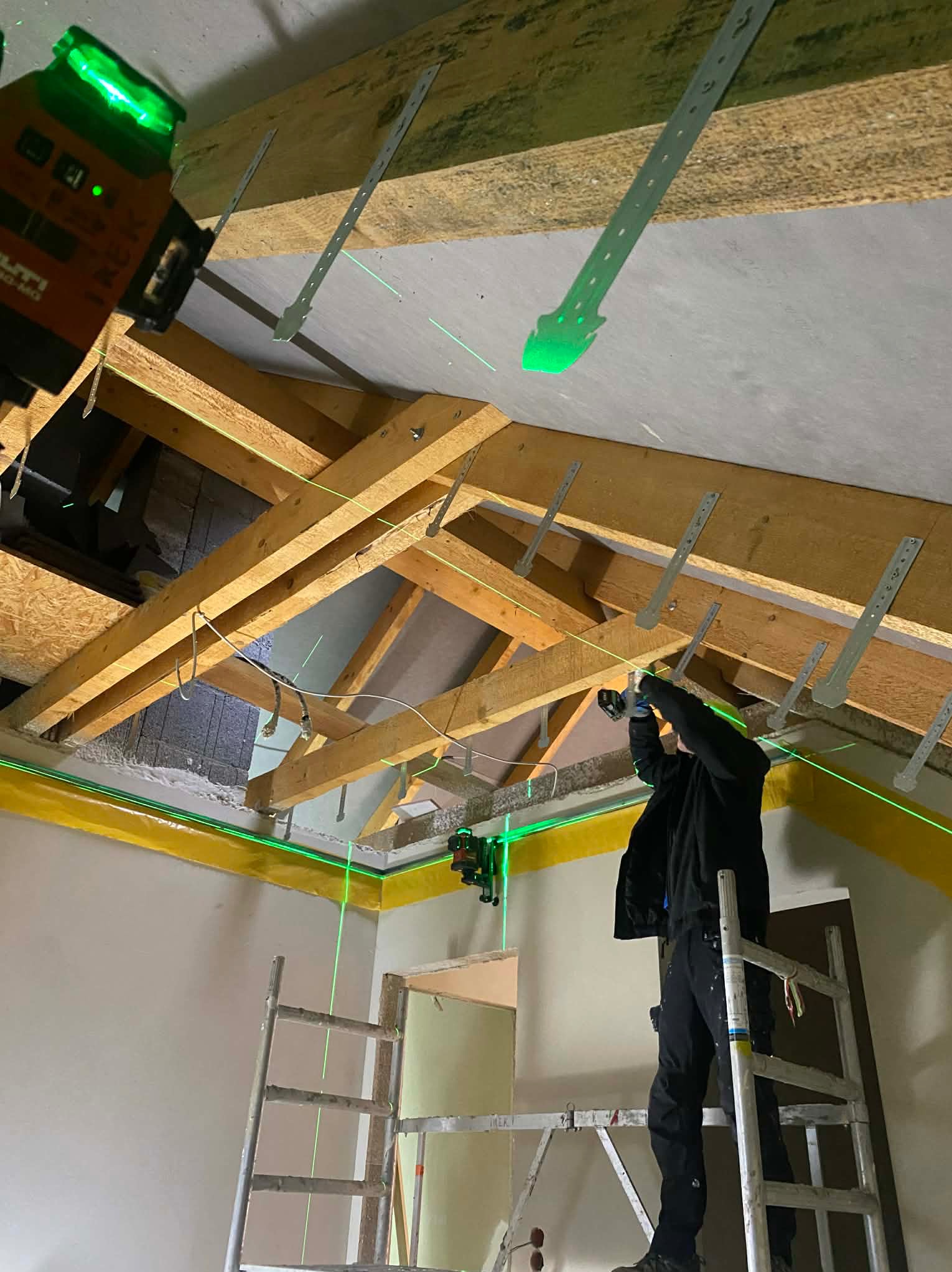 Montaż stelaża pod zabudowę poddasza z użyciem poziomicy laserowej Hilti. Widoczne drewniane krokwie i metalowe wieszaki. Praca na wysokości z drabiny.