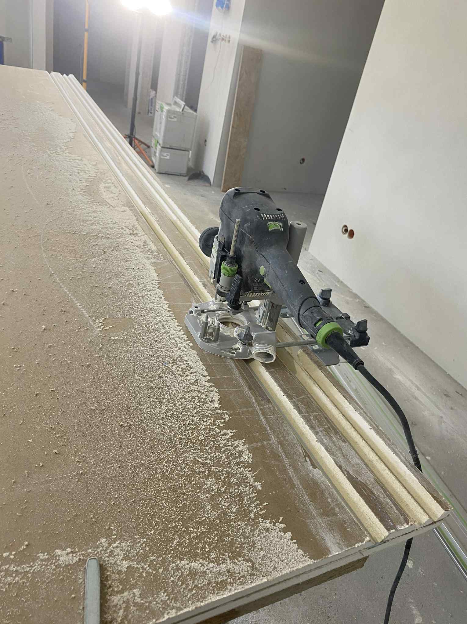 Frezarka krawędziowa Festool w trakcie frezowania płyty gipsowo-kartonowej. Widoczne wióry i prowadnice. Oświetlenie halogenowe w tle. Detal obróbki krawędzi.