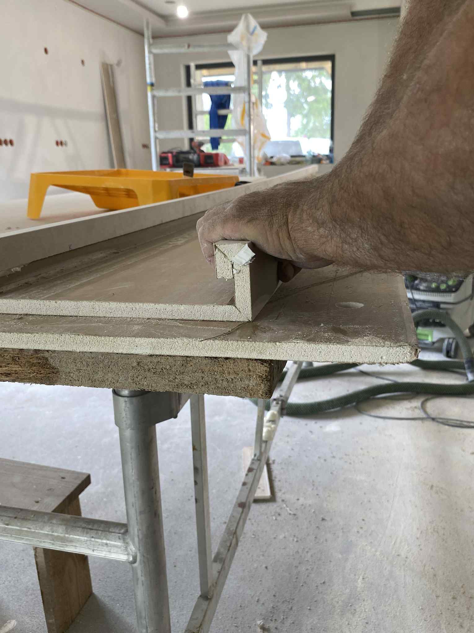 Docinanie płyty gipsowej z profilem w Nowogrodźcu. Widoczna ręka, stół roboczy, drabina i narzędzia. Prace wykończeniowe wewnątrz budynku.