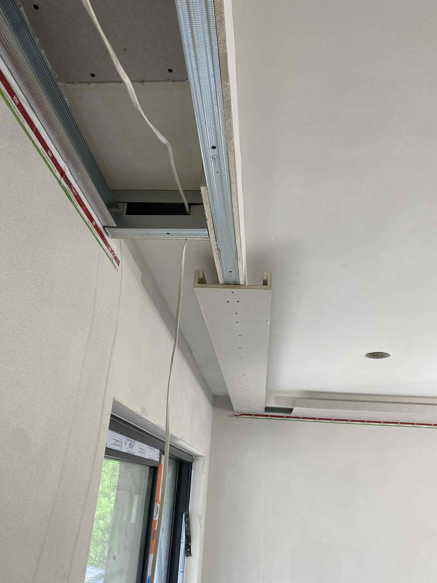 Montaż sufitu podwieszanego z płyt kartonowo-gipsowych na metalowej konstrukcji. Widoczne profile i przewody elektryczne. Świeżo wykończone ściany i okno.