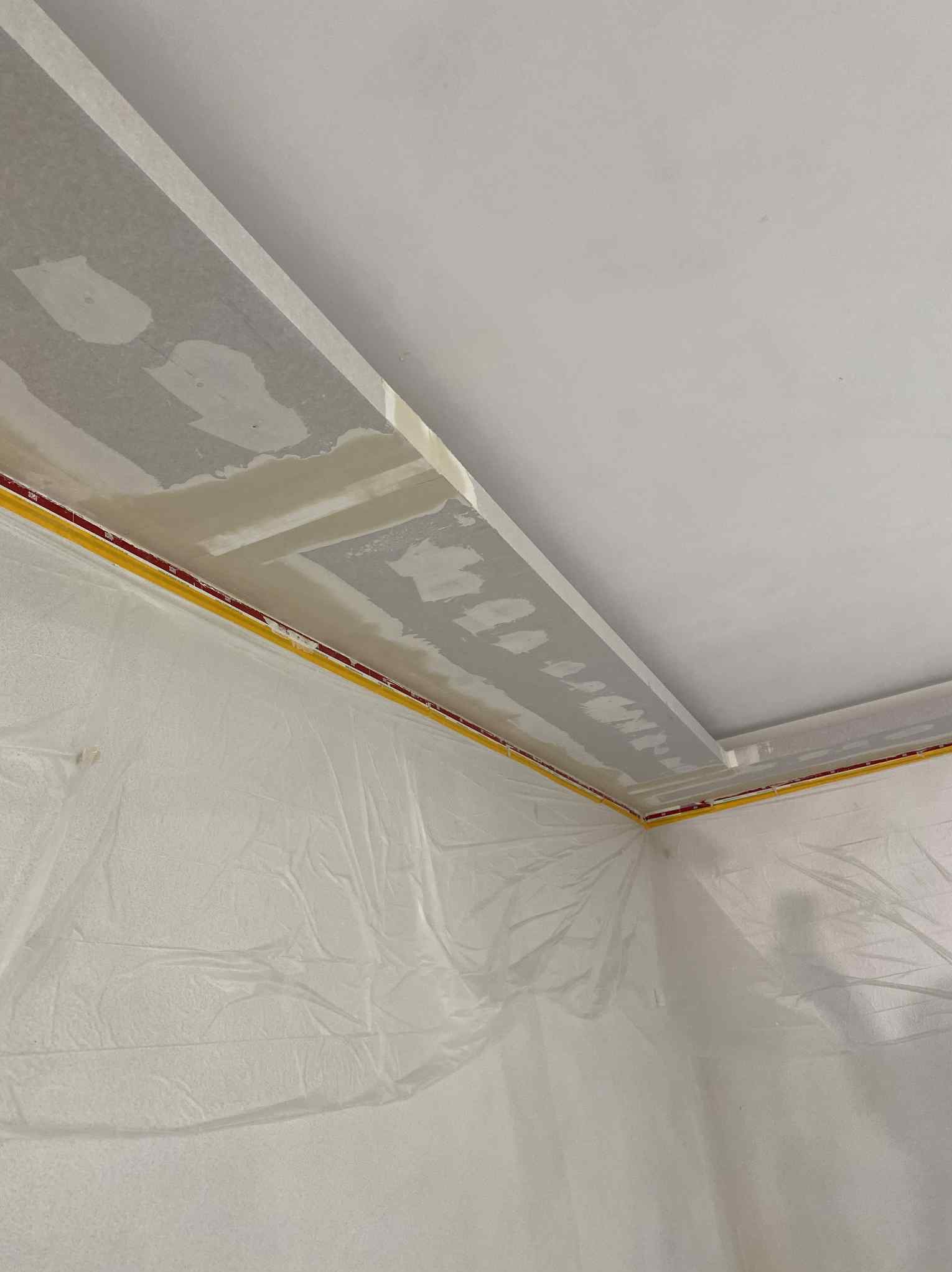 Przygotowanie do szpachlowania narożnika sufitu: zabezpieczona folią ściana, taśma malarska, wstępnie wyszpachlowane łączenie płyt. Widok z dołu, ukośne ujęcie.