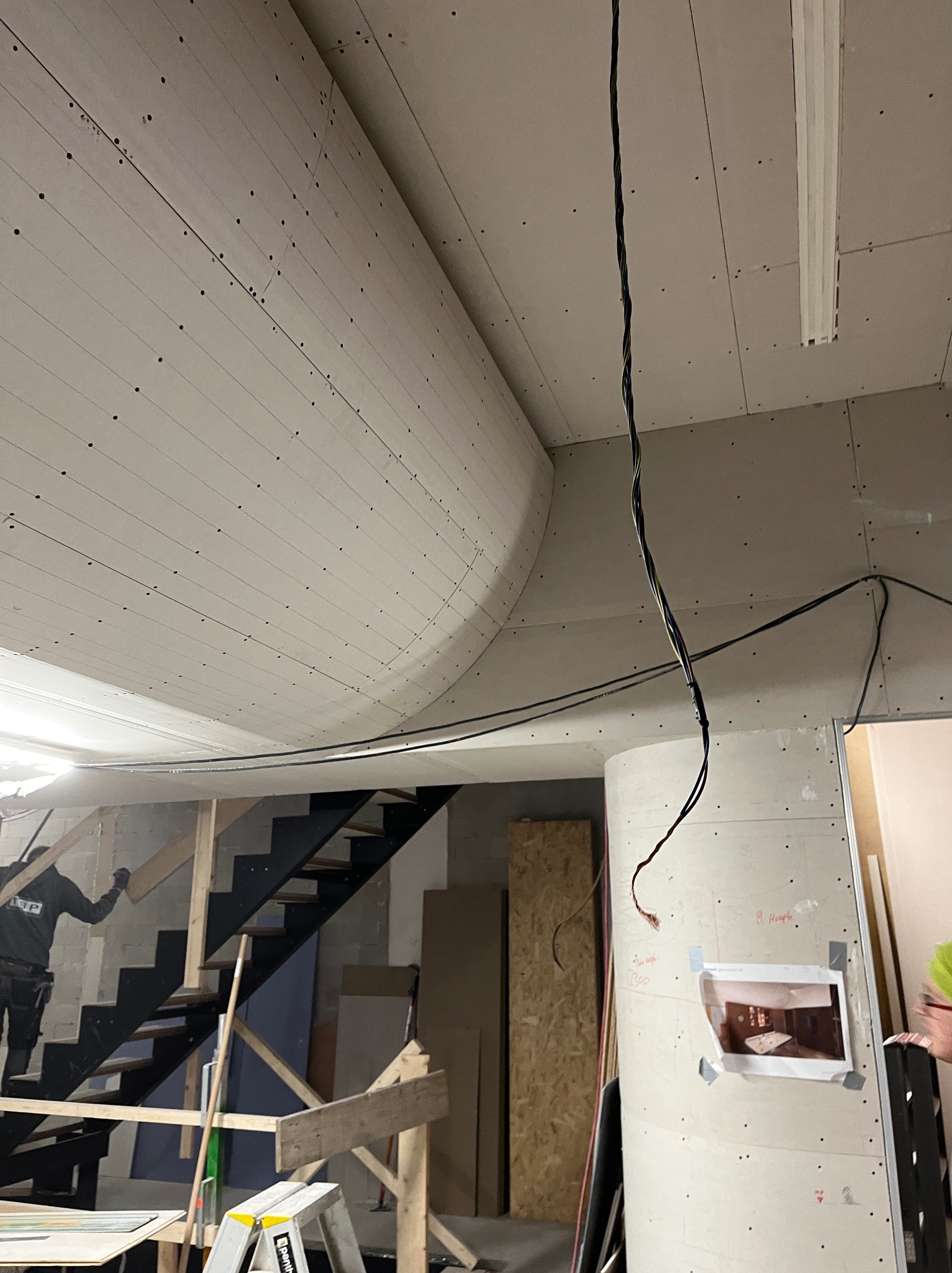 Nietypowy sufit z płyt kartonowo-gipsowych z zaokrąglonym elementem, widoczne kable i fragment schodów. Prace wykończeniowe w trakcie realizacji.