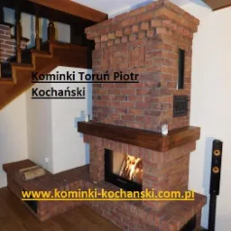 Murowany z cegły kominek z palącym się drewnem, drewniane elementy wykończeniowe, obok schody z drewnianą balustradą.