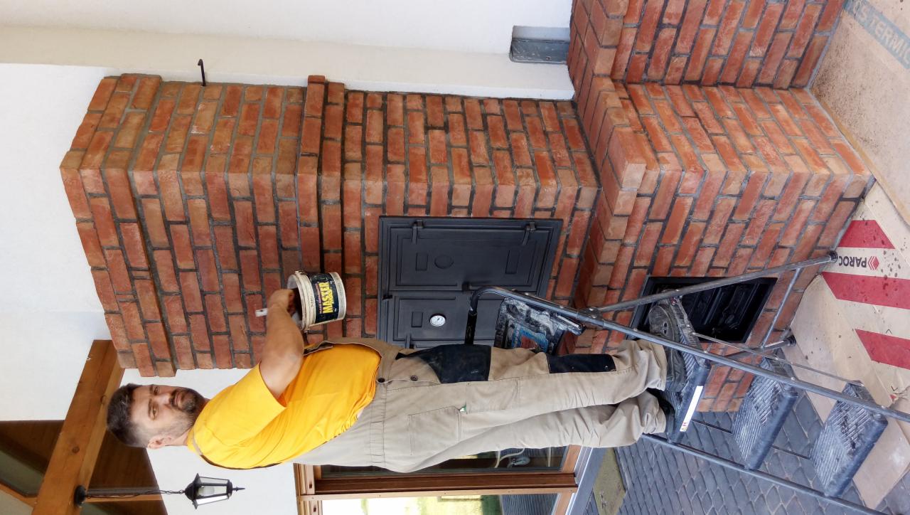 Mężczyzna na drabinie maluje ceglany piec z żeliwnymi drzwiczkami, używając farby z puszki marki Master.