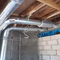 Fragment instalacji wentylacyjnej z rurami aluminiowymi i niebieskimi przewodami elastycznymi, widoczne mocowania do drewnianego stropu, ściana z bloczków betonowych.