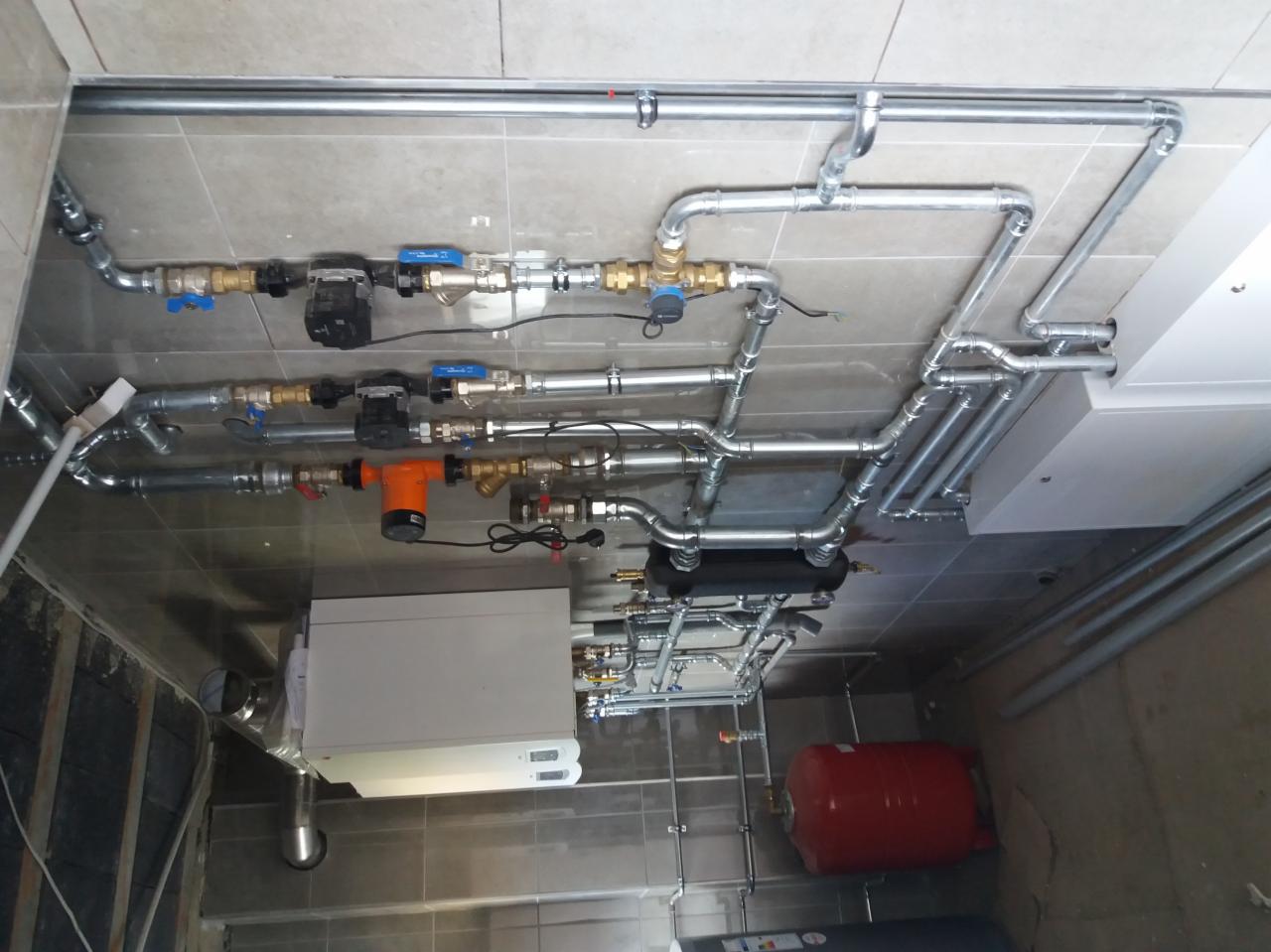 Widok instalacji hydraulicznej i grzewczej pod sufitem z widocznym kotłem, zbiornikiem wyrównawczym, pompą obiegową i licznymi rurami z zaworami.