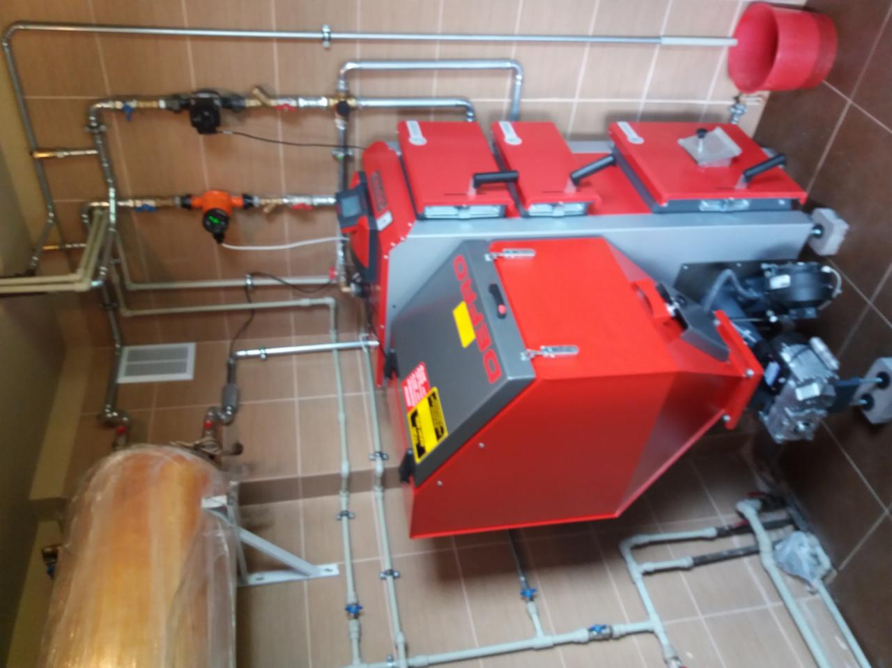 Czerwono-szary piec centralnego ogrzewania marki Defro, z licznymi rurami i zaworami, zamontowany w pomieszczeniu z beżowymi płytkami. Widoczny zbiornik na wodę owinięty folią.