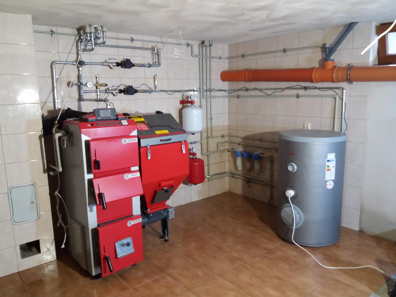 Zainstalowany, czerwono-szary piec na paliwo stałe z podajnikiem, zbiornik buforowy i instalacja hydrauliczna z filtrami w kotłowni wyłożonej płytkami ceramicznymi.
