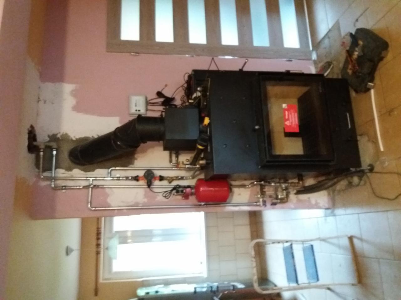 Montaż kotła na pellet z podłączeniami hydraulicznymi i kominowymi w trakcie prac wykończeniowych, widoczny czarny korpus kotła, rury, czerwony zbiornik wyrównawczy, okno i torba narzędziowa w tle.