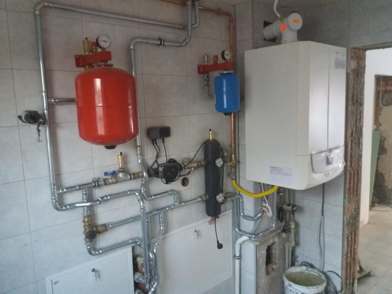 Kompleksowa instalacja grzewcza w kotłowni z widocznym kotłem gazowym, zbiornikiem wyrównawczym w kolorze czerwonym, naczyniem przeponowym w kolorze niebieskim, pompą obiegową i licznymi rurami...