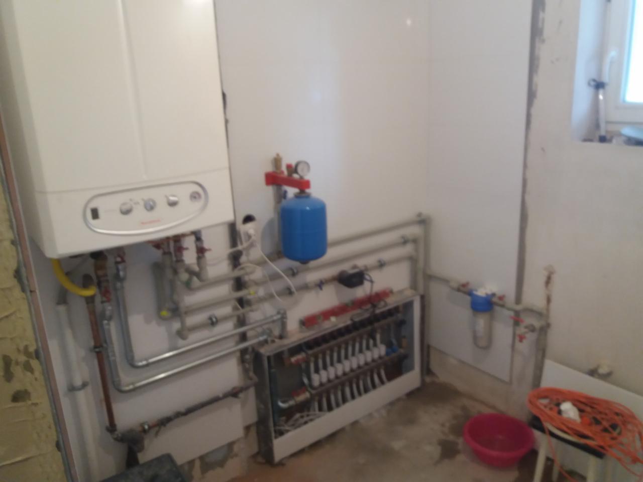 Biały piec gazowy kondensacyjny na ścianie, obok niebieski zbiornik wyrównawczy, szare rury instalacji CO i grzejnikowej, oraz rozdzielacz podłogówki z licznymi pętlami w pomieszczeniu w trakcie...