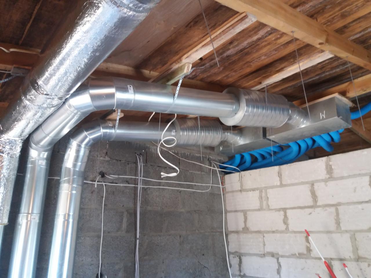 Fragment instalacji wentylacyjnej z rurami aluminiowymi i niebieskimi przewodami elastycznymi, widoczne mocowania do drewnianego stropu, ściana z bloczków betonowych.