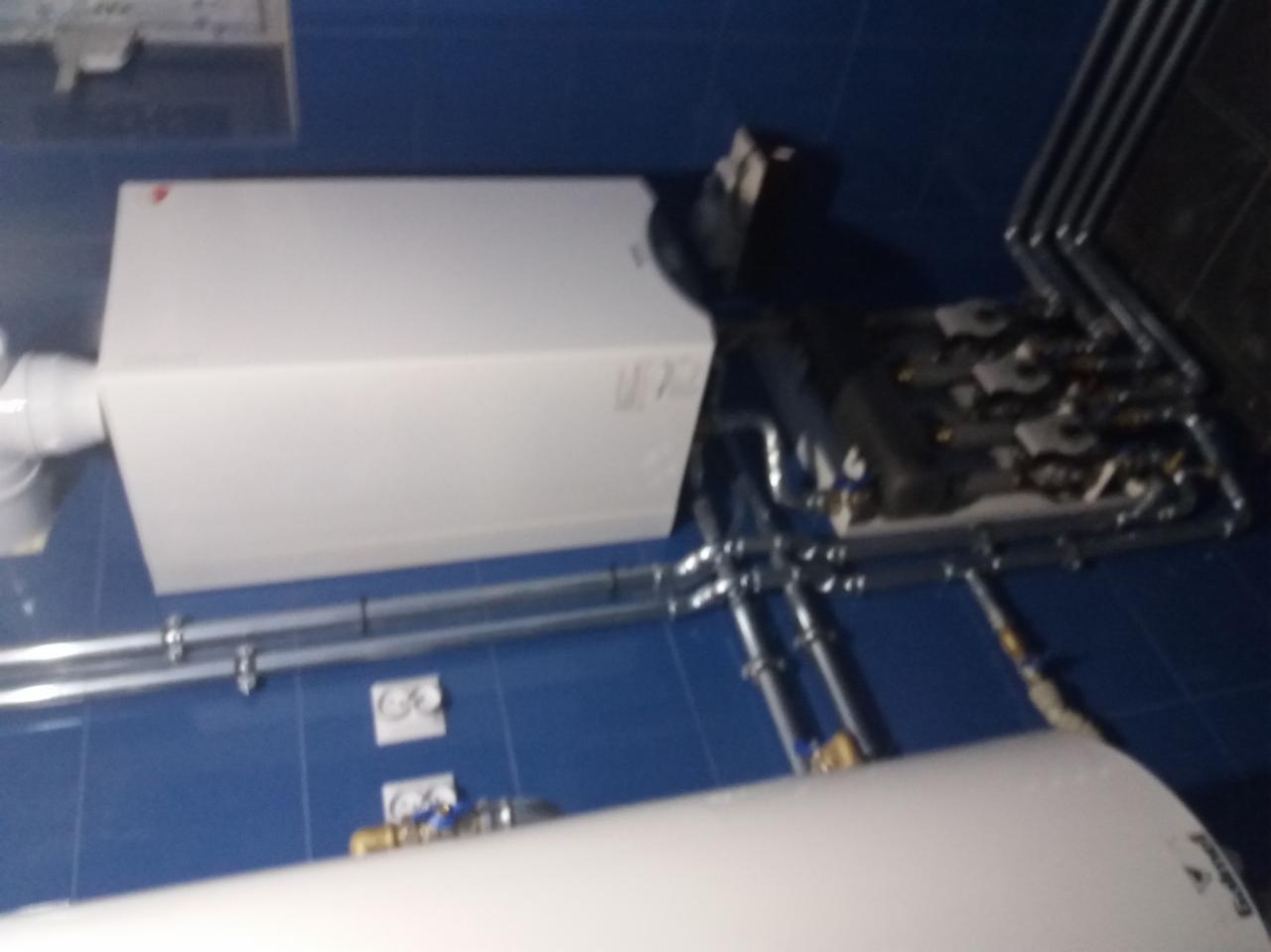 Biały piec grzewczy z podłączonymi rurami instalacji hydraulicznej na tle niebieskich płytek ściennych. Widoczne elementy armatury i złączki.