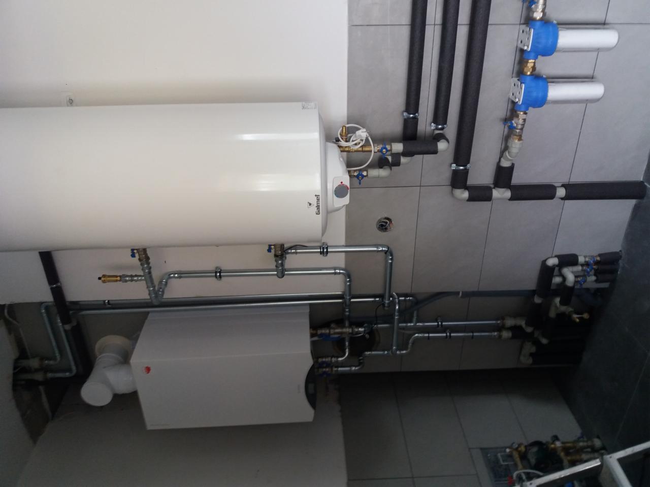 Biały bojler Galmet i rekuperator z szarymi rurami instalacji hydraulicznej, rury wentylacyjne z izolacją termiczną, filtry wody z niebieskimi pokrywami, na tle szarej płytki ceramicznej.