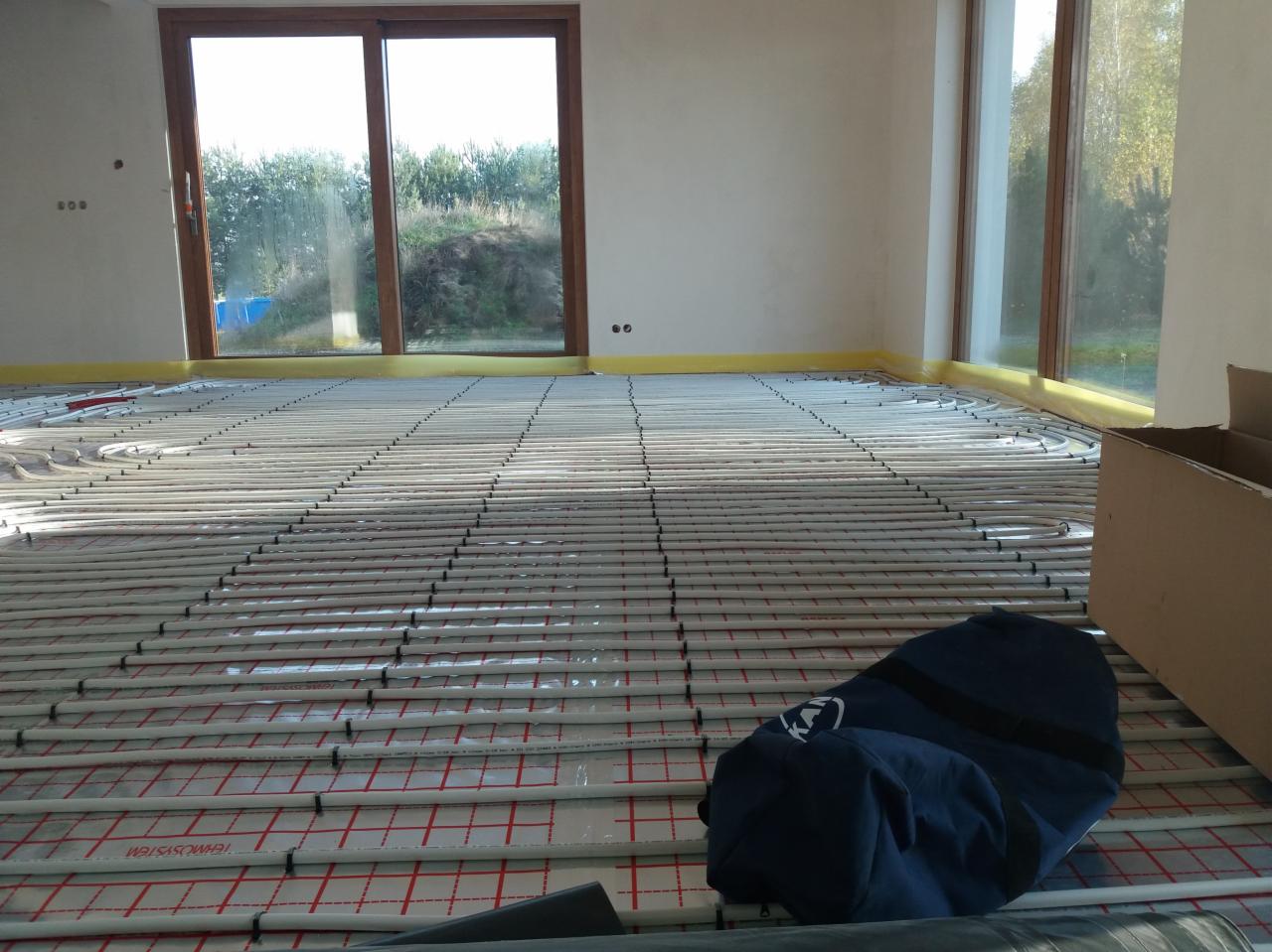 Układanie białych rur grzewczych na podłodze z izolacją w jasnym pomieszczeniu z dużymi oknami, widoczna torba narzędziowa i karton.