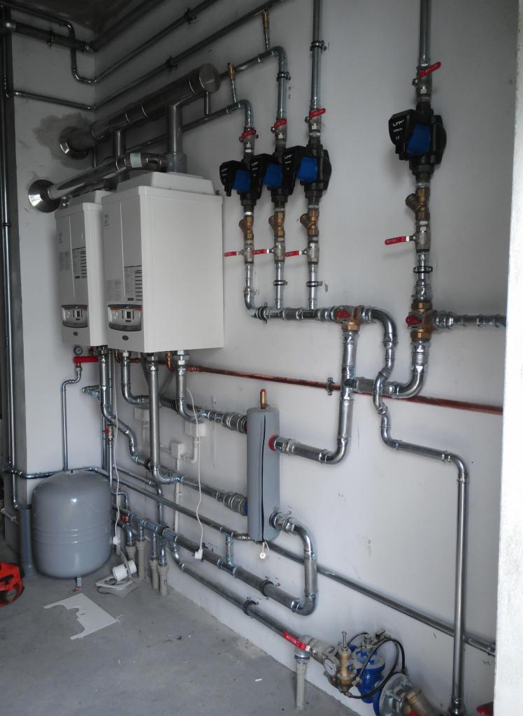 Dwa białe kotły grzewcze zamontowane na ścianie, z licznymi rurami ze stali nierdzewnej, zaworami z czerwonymi uchwytami i pompami obiegowymi z niebieskimi elementami, szary zbiornik wyrównawczy...