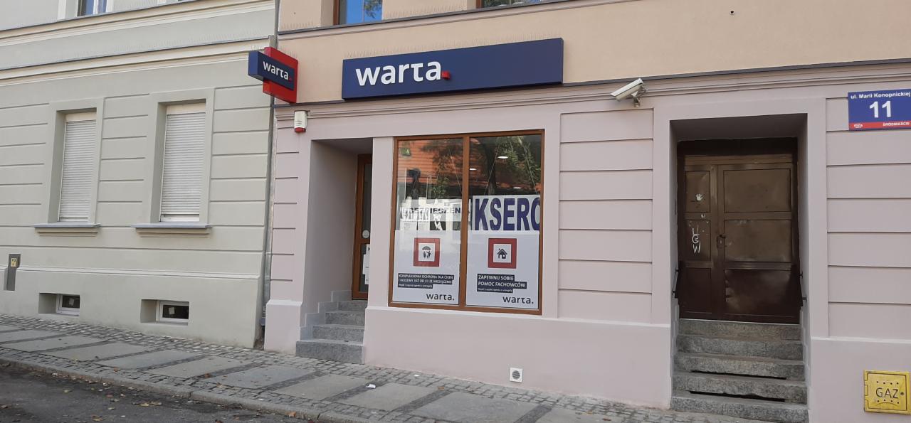 Fasada budynku z szyldami Warta, widoczne okno z naklejonymi plakatami informacyjnymi oraz tabliczka z adresem ul. Marii Konopnickiej 11.
